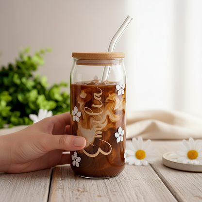 Ein personalisiertes Glas mit Bambusdeckel und Glasstrohhalm, gefüllt mit Eiskaffee, in dem sich Milch dekorativ verteilt. Auf dem Glas sind weiße Blumen und der Schriftzug „Emilia“ zu sehen. Eine Hand hält das Glas auf einem Holztisch; im Hintergrund befinden sich unscharfe grüne Pflanzen und Gänseblümchen.