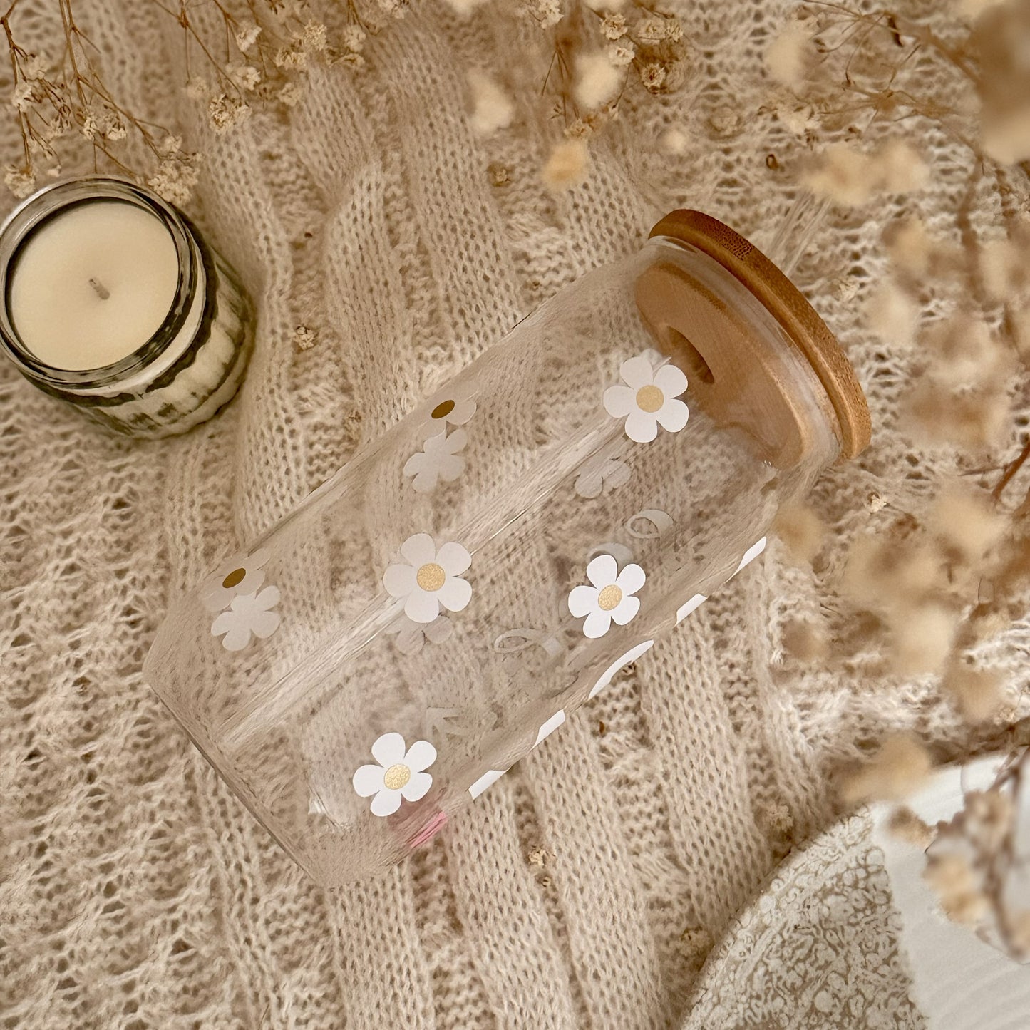 Trinkglas mit verspieltem Gänseblümchen-Muster in Weiß und Gold und Bambusdeckel, auf heller Strickdecke mit Kerze und Trockenblumen – Blumenglas im Coquette-Look.