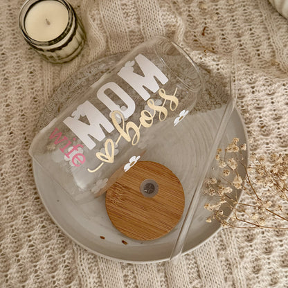Trinkglas „MOM wife boss" in Weiß, Rosa und Gold auf hellem Teller mit Bambusdeckel und Gänseblümchen-Muster, dekoriert mit Kerze und Trockenblumen – Geschenk für Mamas zum Kaffee.