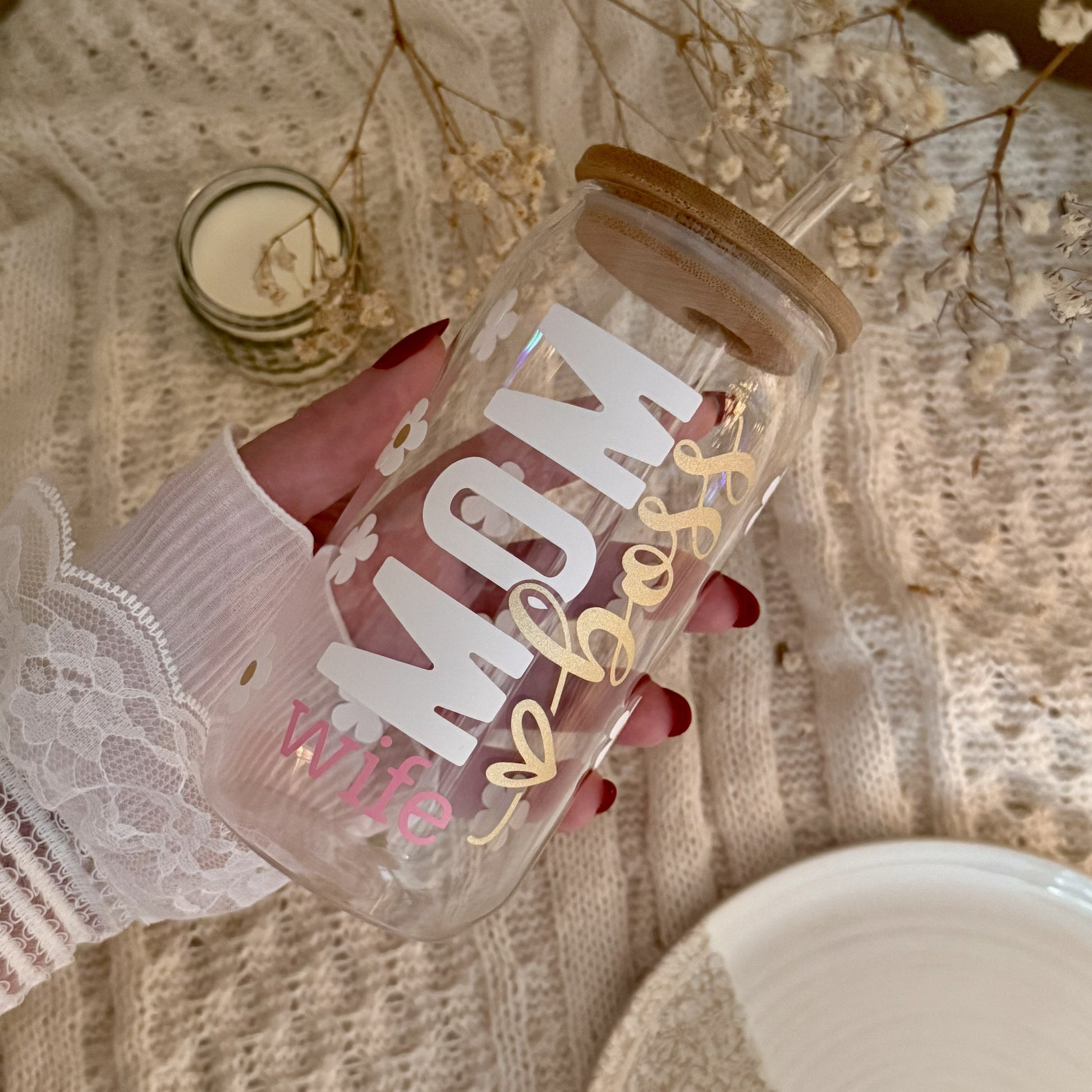 Trinkglas „MOM wife boss" aus Klarglas in der Hand gehalten, Schriftzug in Weiß, Rosa und Gold, mit Bambusdeckel und Gänseblümchen – Geschenkidee für Mamas.